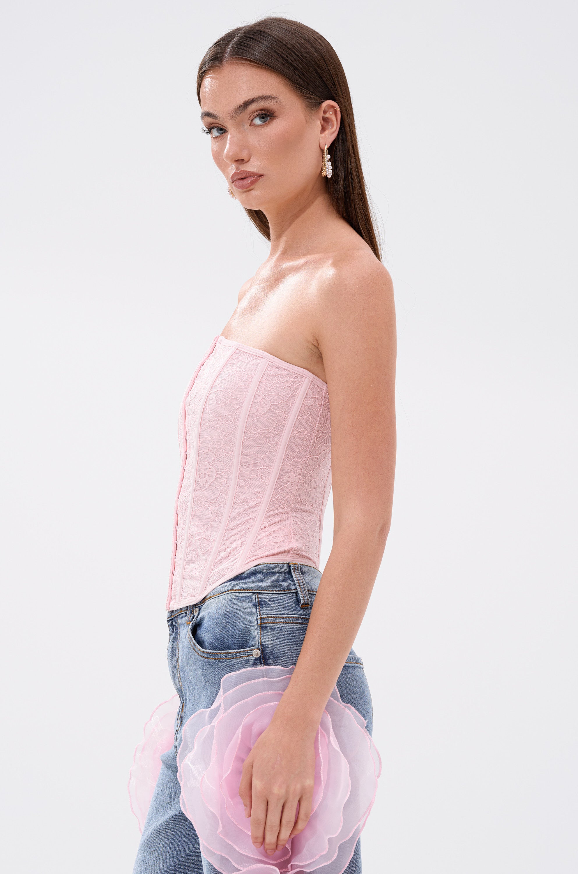 A woman with long brown hair wears the FAIRY TALE CORSET TOP in light pink and blue jeans, holding a pink flower-shaped accessory, standing against a plain white background.