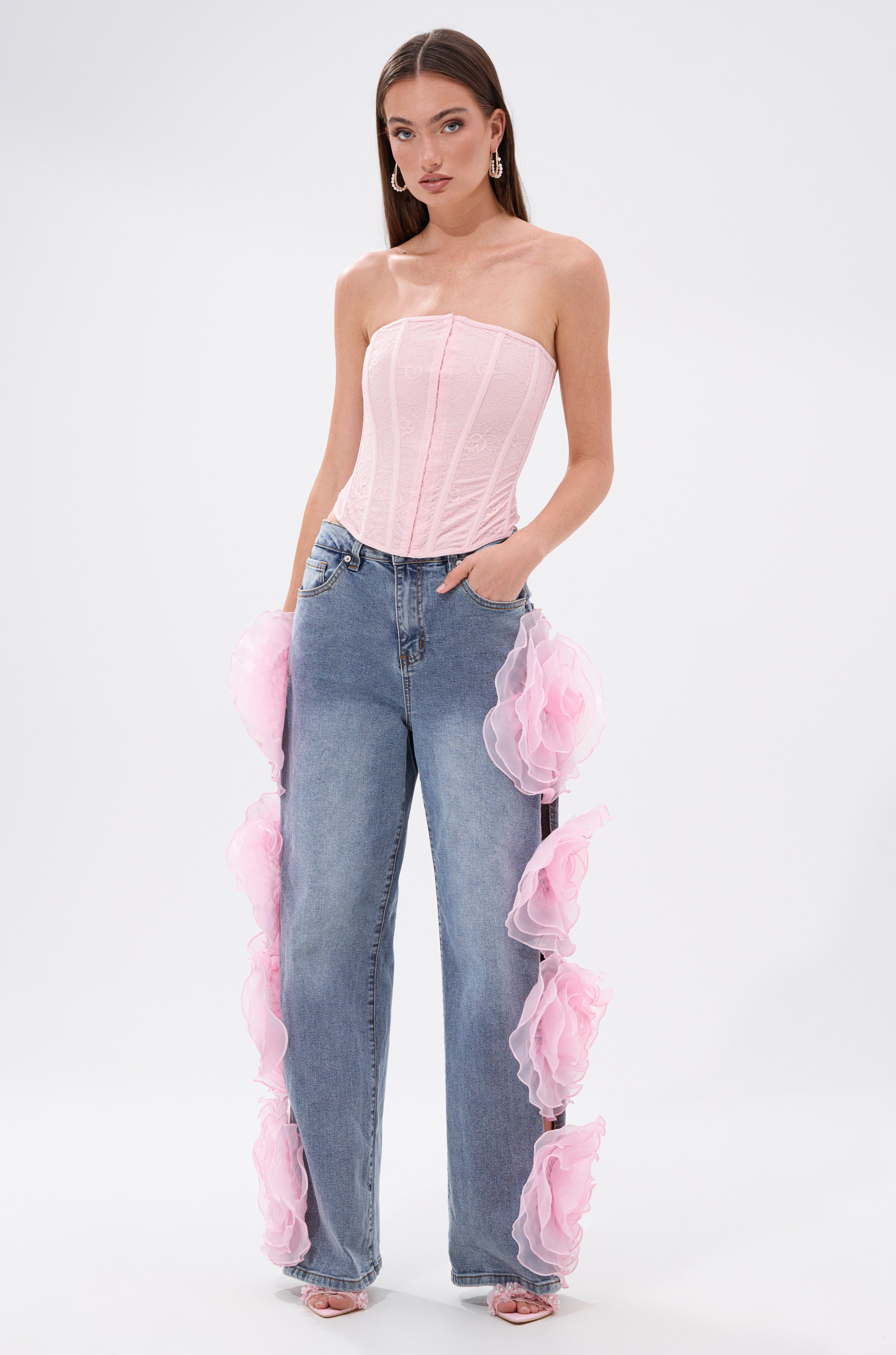 A woman models the FAIRY TALE CORSET TOP, a strapless piece with floral lace details, paired with high-waisted blue jeans featuring large pink ruffled side accents, posing confidently against a plain white background.