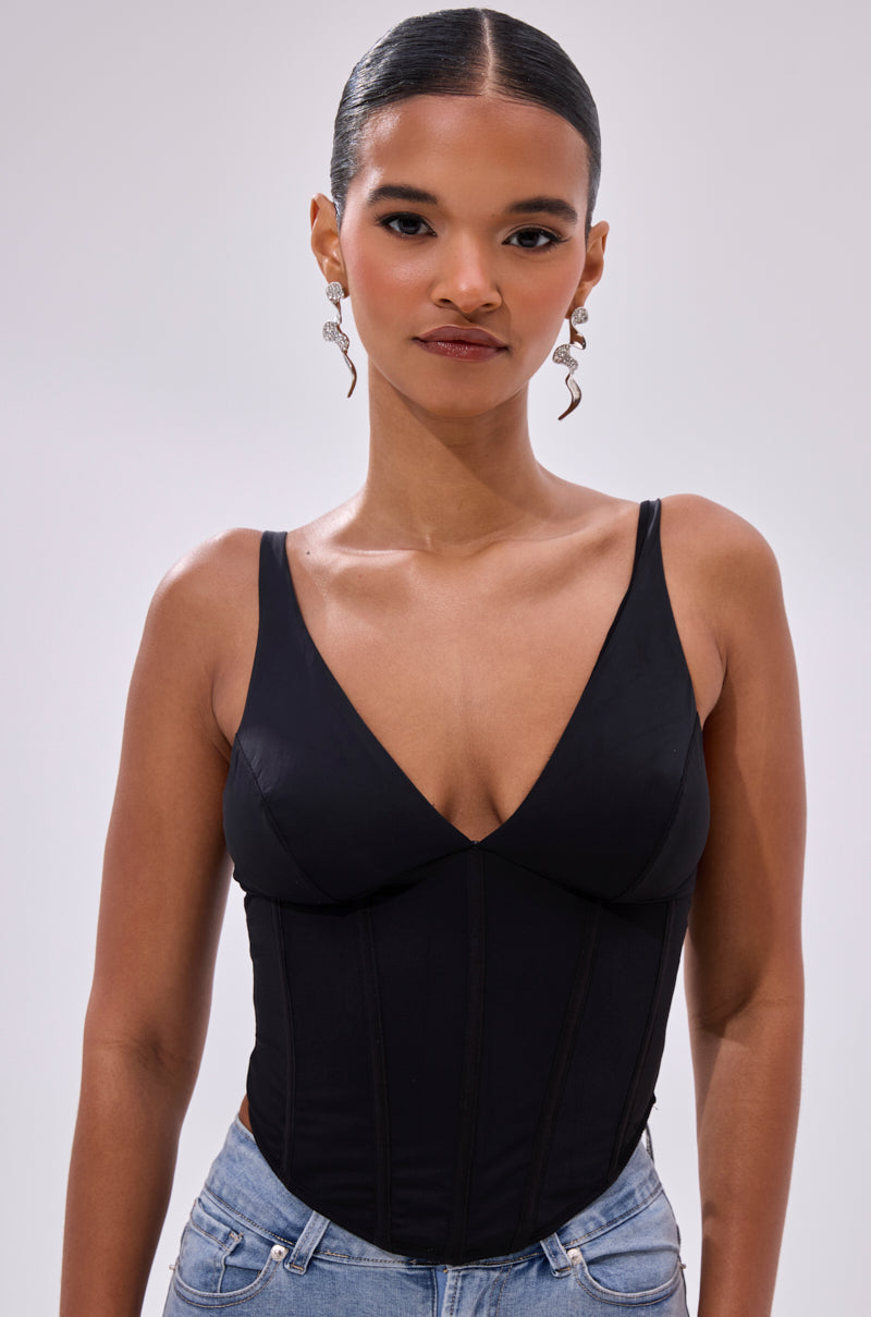 A woman with sleek, pulled-back hair and dangly earrings wears the FREE SPIRIT SLEEVELESS CORSET TOP IN BLACK styled with light blue jeans, standing against a plain light background.