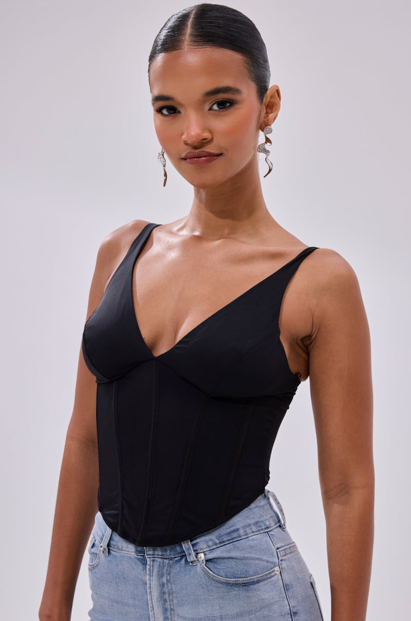 A woman with sleek dark hair and subtle makeup stands confidently against a plain light background, wearing the AKIRA FREE SPIRIT SLEEVELESS CORSET TOP IN BLACK with a deep V-neck, light blue jeans, and dangling earrings.