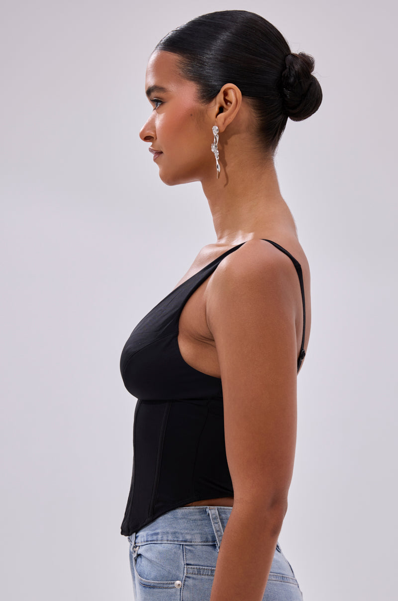 A woman with her hair in a sleek bun, silver drop earrings, and light blue jeans stands in profile against a neutral background, wearing the FREE SPIRIT SLEEVELESS CORSET TOP IN BLACK.