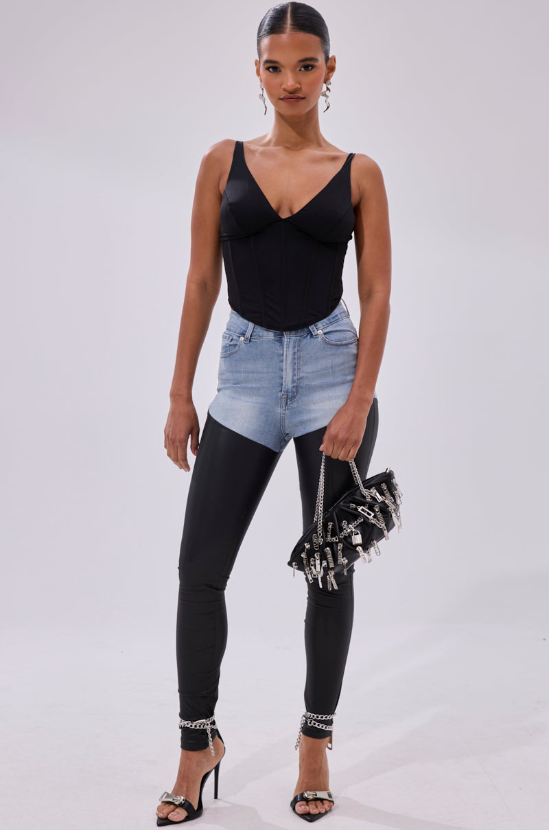 A woman stands confidently in the FREE SPIRIT SLEEVELESS CORSET TOP IN BLACK, paired with light blue high-waisted denim shorts over black leggings, black heels, a black studded handbag, sleek hair, and statement earrings.