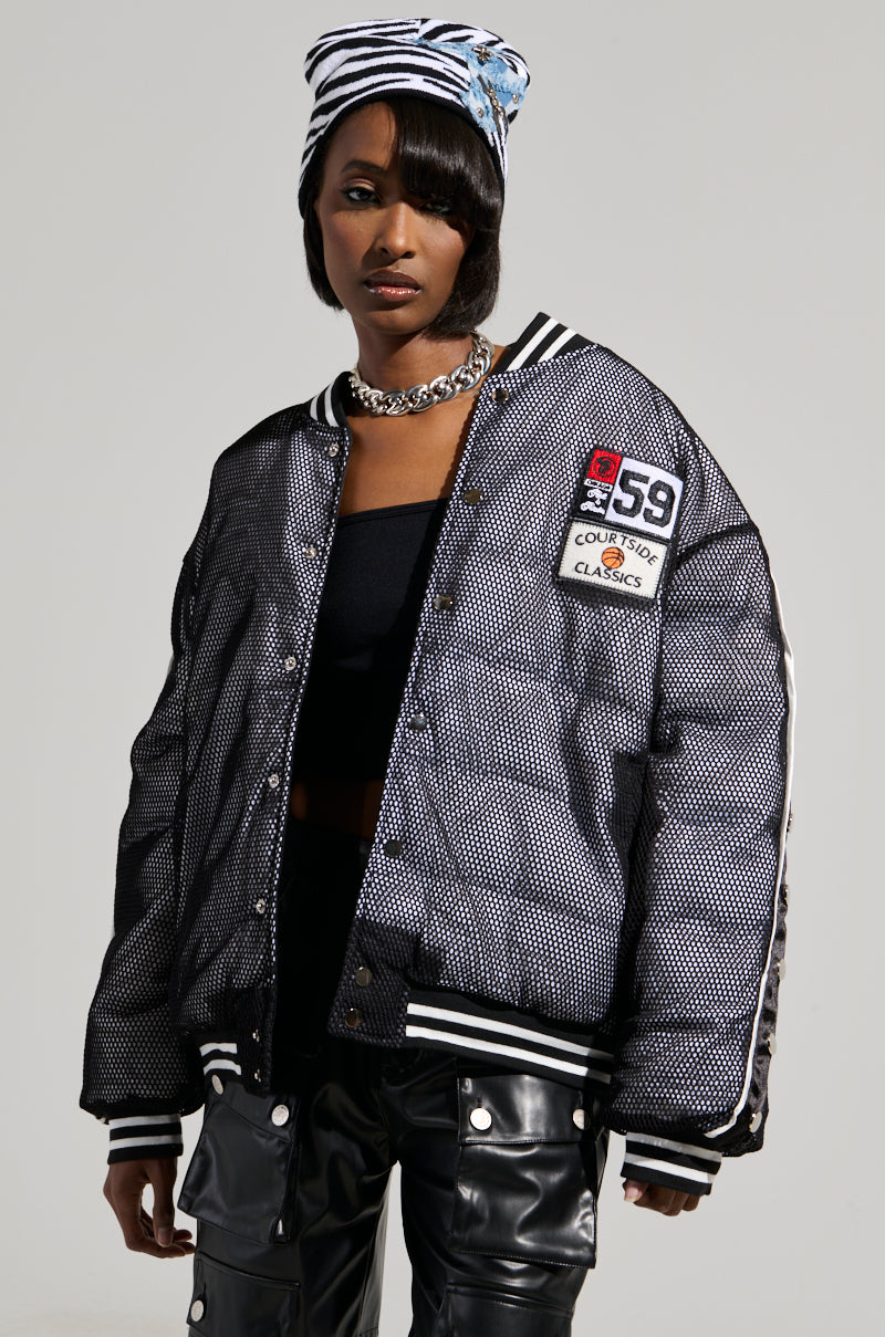 Sporting the PLAYERS ONLY JERSEY BOMBER, a person exudes confidence with its black and white patterned design, complemented by athletic patches. The jacket is paired with a black top and accessorized with a silver chain necklace and a blue-and-white designed beanie, creating an effortlessly cool vibe. Black pants finish off the ensemble against a plain background.