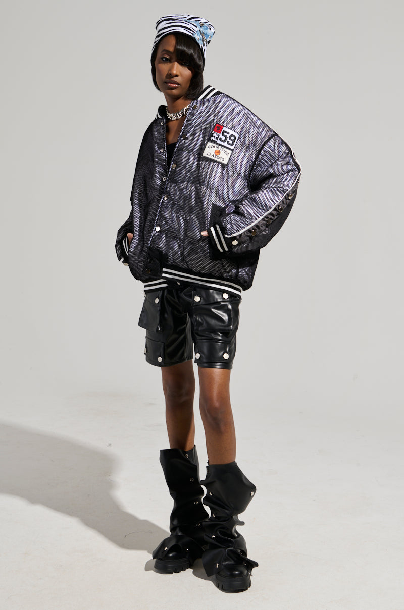 A person wearing the PLAYERS ONLY JERSEY BOMBER in black and silver with textured designs and athletic patches pairs it confidently with black leather shorts and boots. A striped beanie and chain necklace perfectly complement the edgy ensemble against a plain, neutral background.