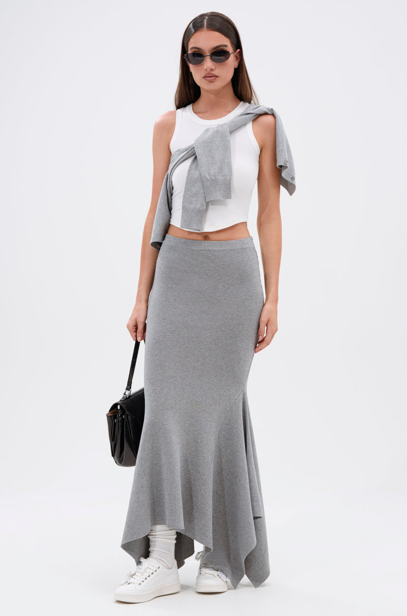 Woman wearing sunglasses, a white sleeveless crop top, gray sweater draped over her shoulders, the TAKE IT BACK KNIT MAXI SKIRT with an asymmetrical hem, white socks, white sneakers, and carrying a black handbag.