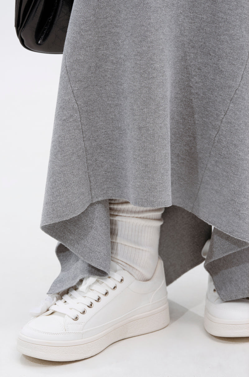 A close-up of a person wearing the TAKE IT BACK KNIT MAXI SKIRT in gray, paired with white socks and sneakers. A partially visible black bag appears at the top left against a plain light background.