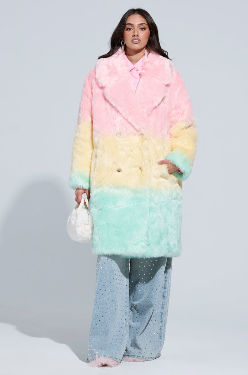 Dressed confidently in a pastel rainbow SORBET FAUX FUR TRENCH, the person with long hair carries a small white handbag. Their ensemble is completed with wide-legged jeans adorned with pearl embellishments, set against a simple, light background.