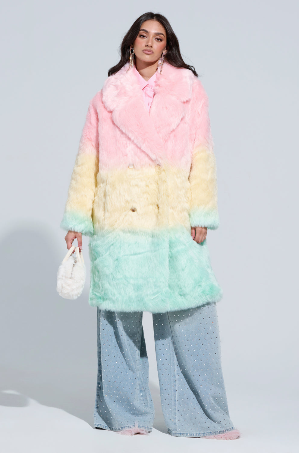 A woman in a SORBET FAUX FUR TRENCH, featuring an oversized pastel rainbow design, stands against a light background. The ombre colorway of her coat highlights pink, yellow, and teal stripes. She pairs it with wide-leg light blue pants adorned with small embellishments and carries a white textured handbag.