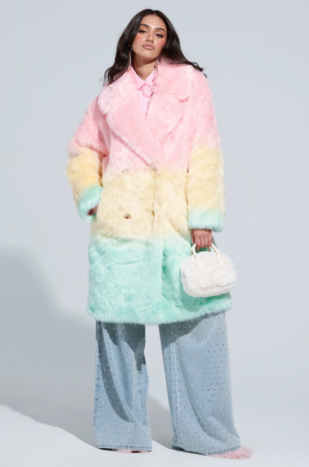 A person wearing the SORBET FAUX FUR TRENCH in an oversized ombre of pink, yellow, and mint is paired with wide-legged blue jeans embellished with small silver studs. They carry a fuzzy white handbag and have long, dark hair.