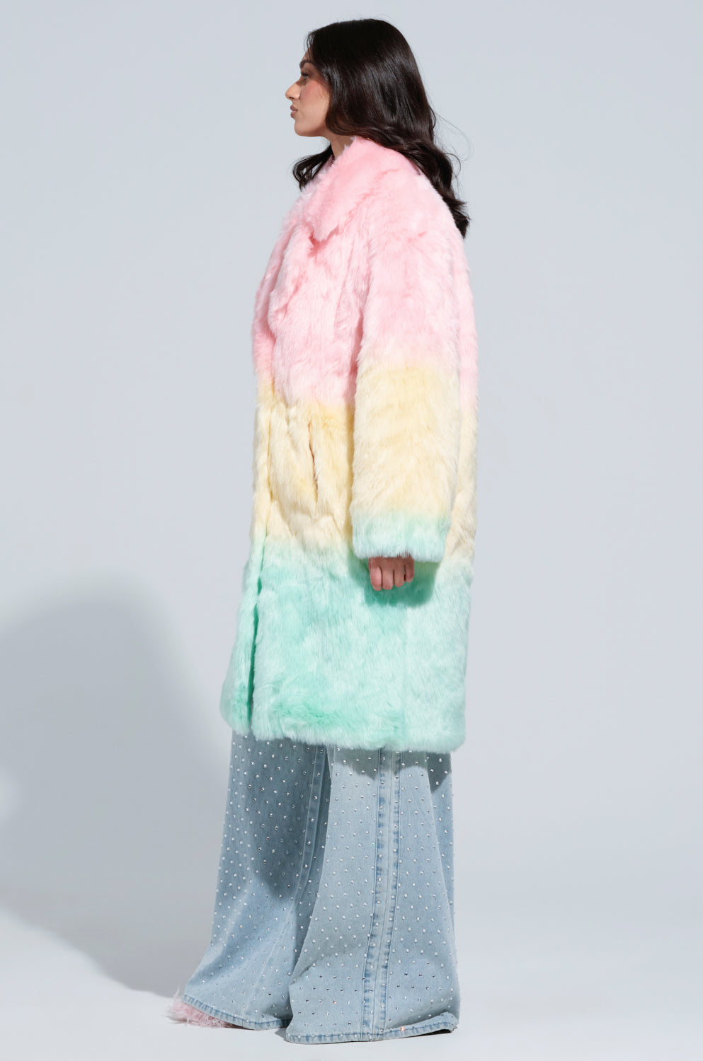 A person with long dark hair is wearing the pastel Sorbet Faux Fur Trench in shades of pink, yellow, and green. They have paired it with wide-legged jeans featuring white dots. This fashionable individual stands sideways against a plain light gray background, exuding an effortlessly chic appearance.