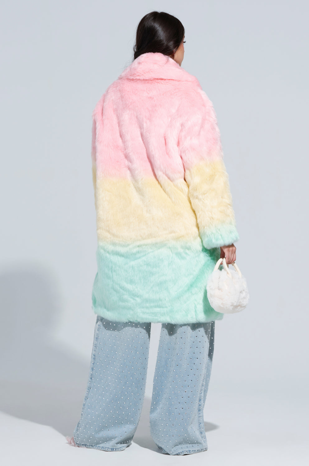 A person with long hair stands facing away, clad in the SORBET FAUX FUR TRENCH featuring pink, yellow, and mint green shades. They carry a small fluffy white bag and sport wide-legged jeans adorned with silver studs against a plain backdrop, elegantly highlighting their pastel accessories.