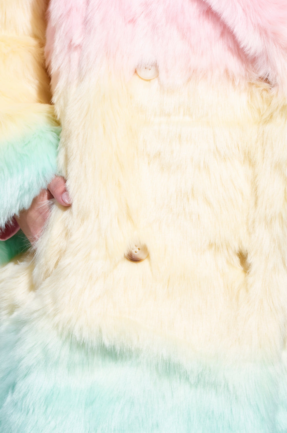 A close-up shot of the SORBET FAUX FUR TRENCH highlights its oversized design, featuring beautiful shades of yellow and pink. The large buttons enhance its charm, while a hand partially appears on the left, perfectly accentuating the vibrant look with pastel accessories.