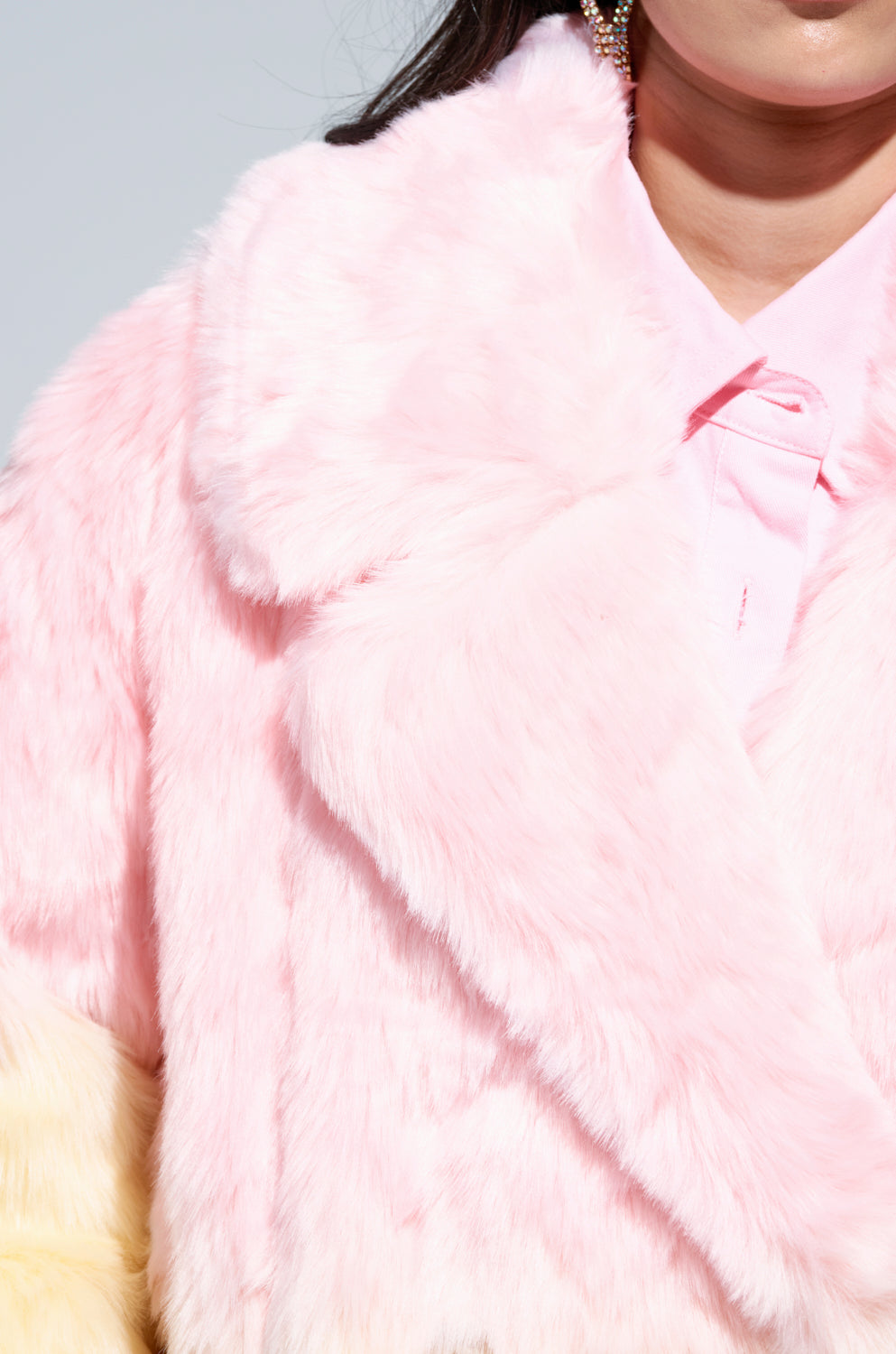 A close-up shows someone wrapped in the oversized SORBET FAUX FUR TRENCH, layered over a matching pink shirt. While their head is mostly out of frame, a dangling earring can be partially seen. The fabric appears soft and plush in texture.
