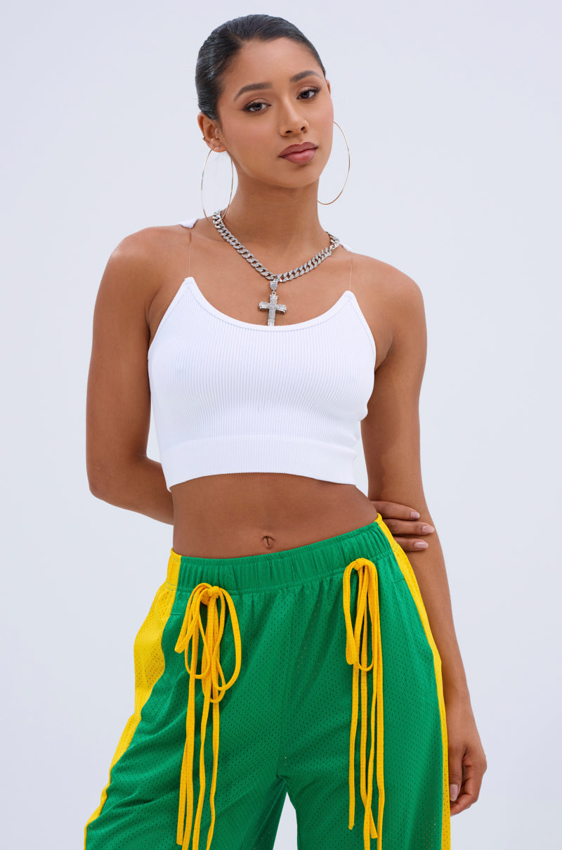 A woman poses against a plain background, wearing the INVISIBLE STRAP CROPPED TANK TOP, green and yellow track pants with yellow drawstrings, large hoop earrings, and a chunky chain necklace with a cross pendant.