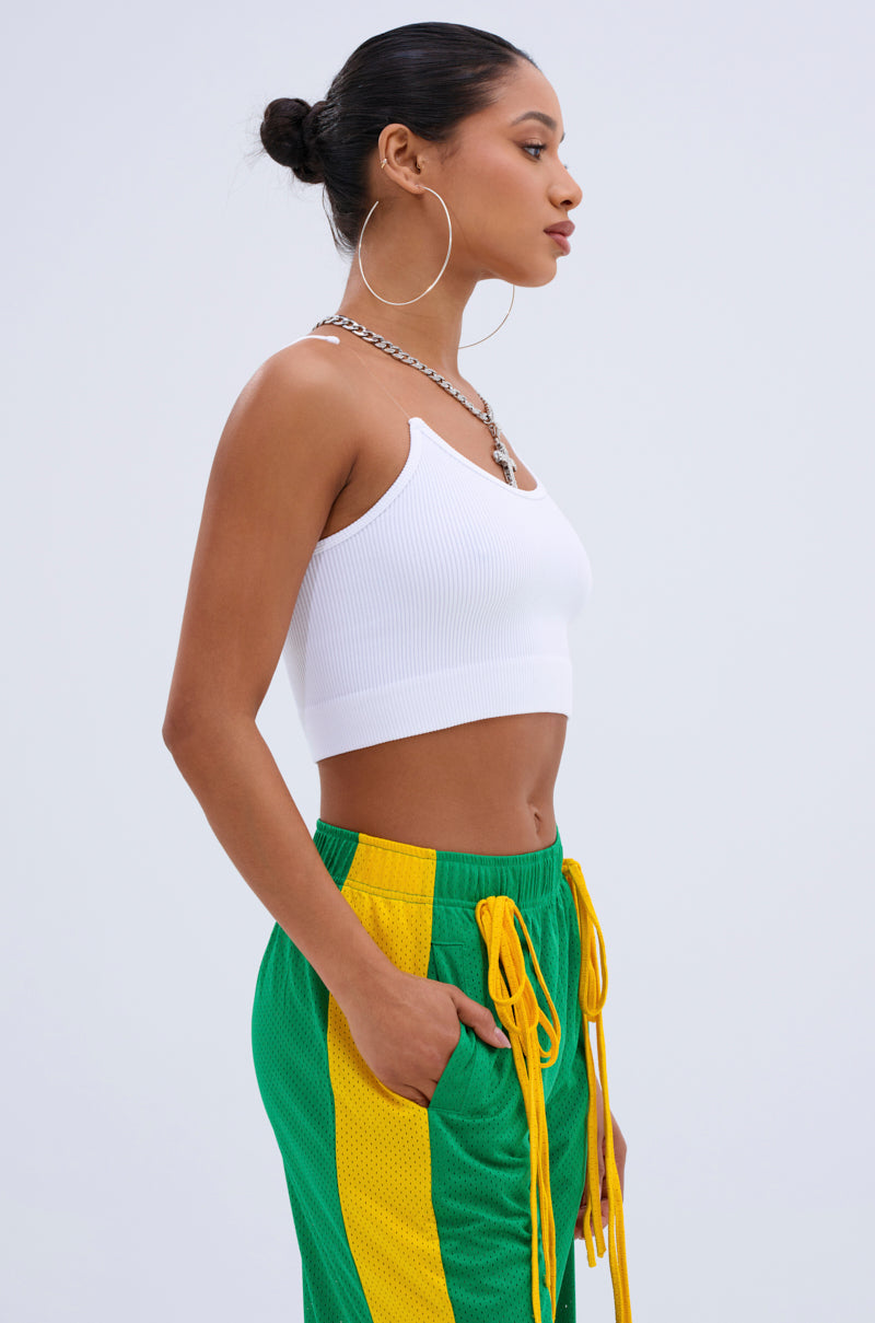 A woman in profile against a plain background wears the INVISIBLE STRAP CROPPED TANK TOP in white with green and yellow athletic shorts, large hoop earrings, a silver chain necklace, and her hair styled in a neat bun.