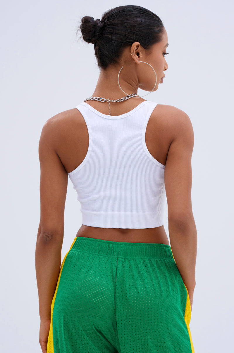 A woman with her hair in a bun is seen from behind, wearing the INVISIBLE STRAP CROPPED TANK TOP, green athletic shorts with yellow side stripes, large hoop earrings, and a silver chain necklace.