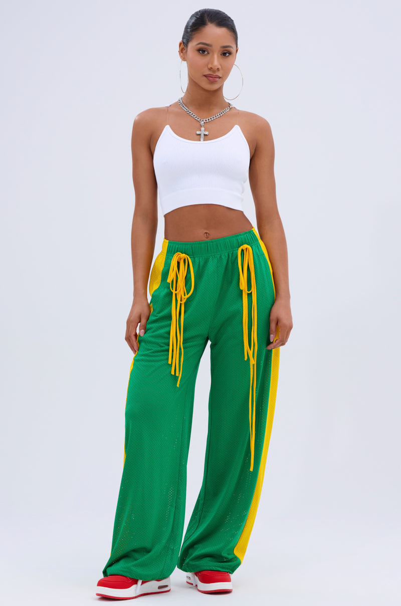 A woman wears the INVISIBLE STRAP CROPPED TANK TOP in white, styled with green wide-leg pants featuring yellow side stripes and ties, red and white sneakers, a necklace, and earrings against a plain light background.