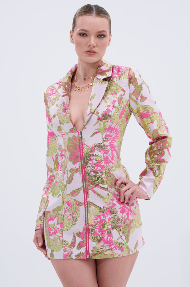 A woman poses confidently in the PRETTY IN PINK BROCADE BLAZER DRESS, a metallic floral piece with pink, green, and gold hues. It features a deep V-neckline and front zipper, styled with a gold necklace and slicked-back hair for a chic look.