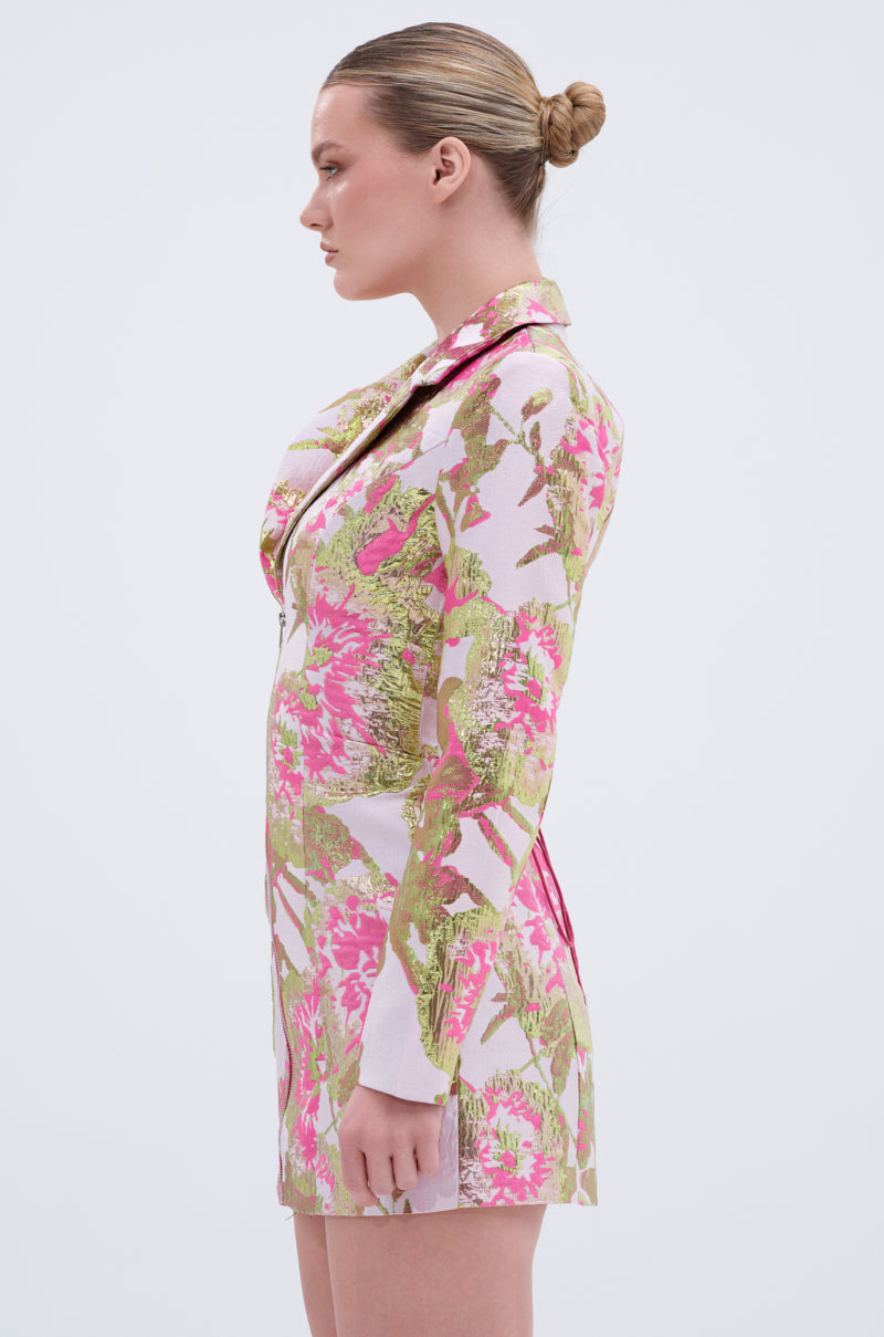 A woman with blonde hair in a bun stands in profile, wearing the PRETTY IN PINK BROCADE BLAZER DRESS featuring a floral pattern in pink, green, and white against a plain light background.