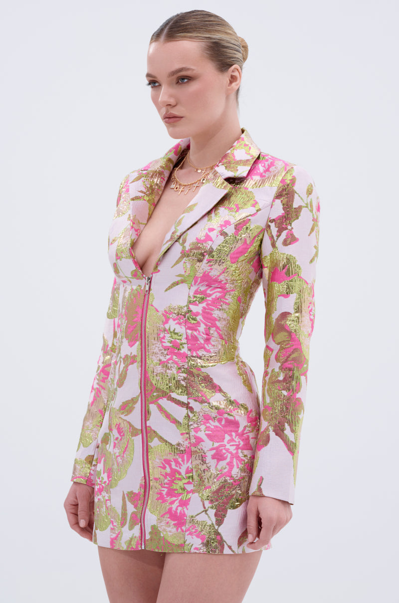 A woman wears the PRETTY IN PINK BROCADE BLAZER DRESS, featuring metallic pink, green, and gold floral patterns. She stands against a plain light background with her hair pulled back and accessorizes with a gold necklace.