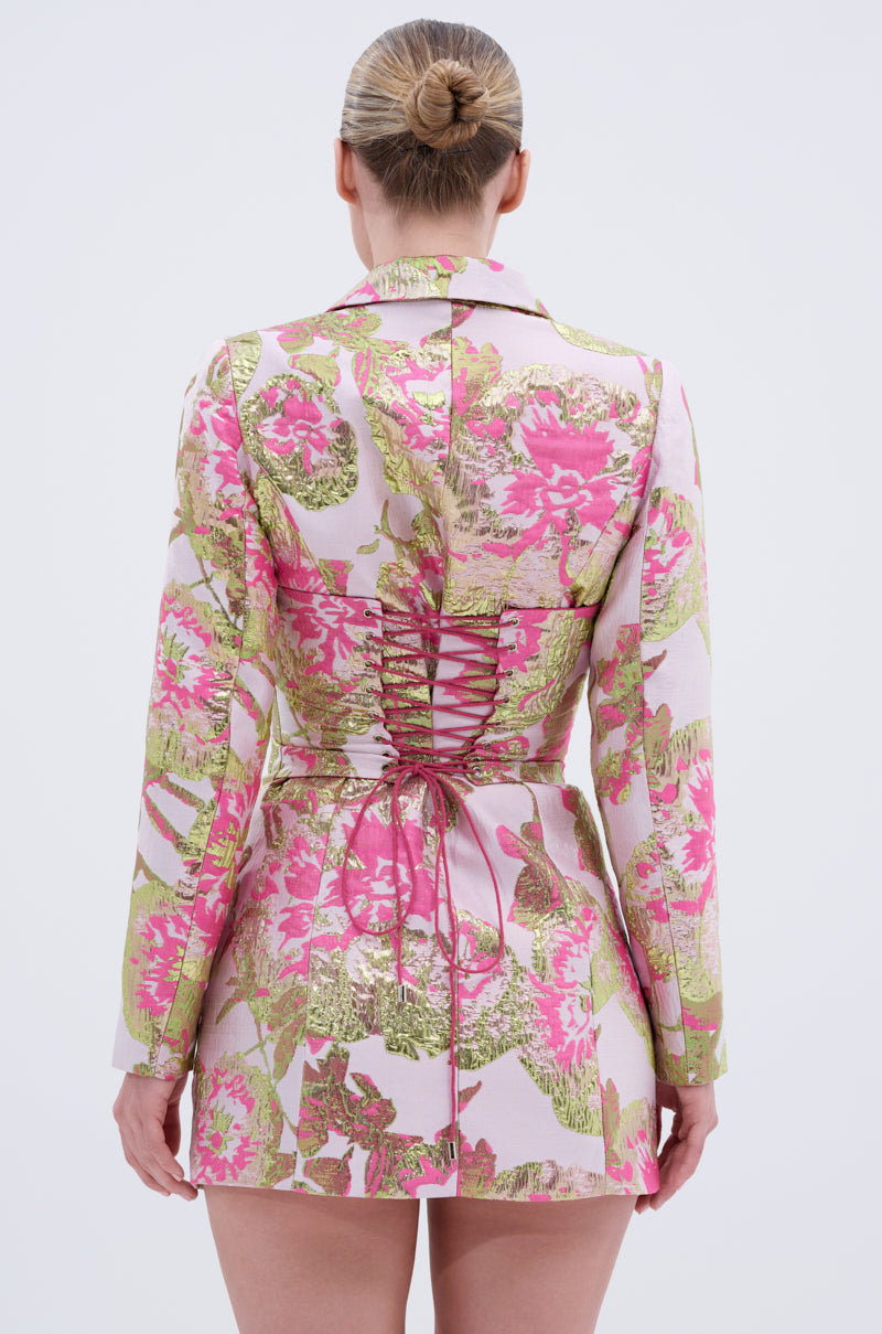 A woman with her hair in a neat bun is seen from behind, wearing the PRETTY IN PINK BROCADE BLAZER DRESS featuring a metallic floral pattern and a corset-style lace-up back.