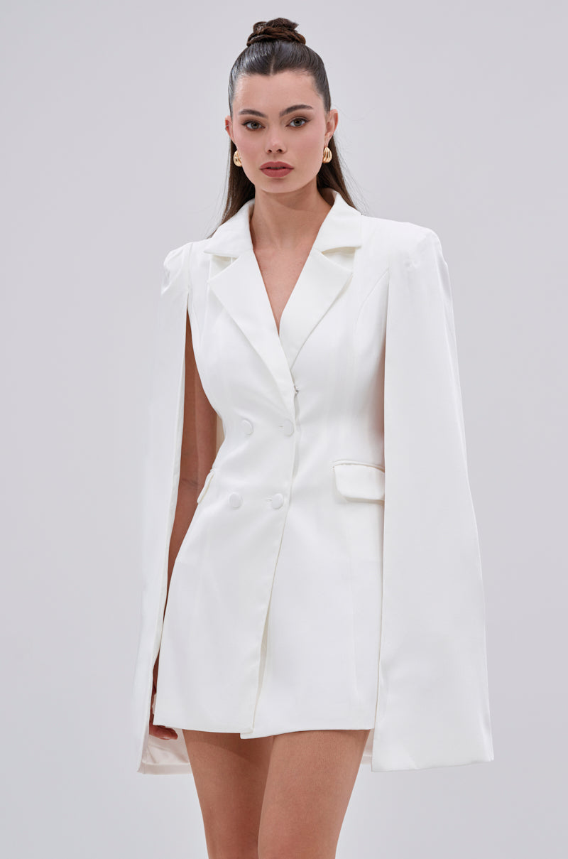 A woman with long brown hair in a high bun wears the A LA MODE MINI BLAZER DRESS IN WHITE with cape sleeves, standing against a plain light background and looking confidently at the camera.