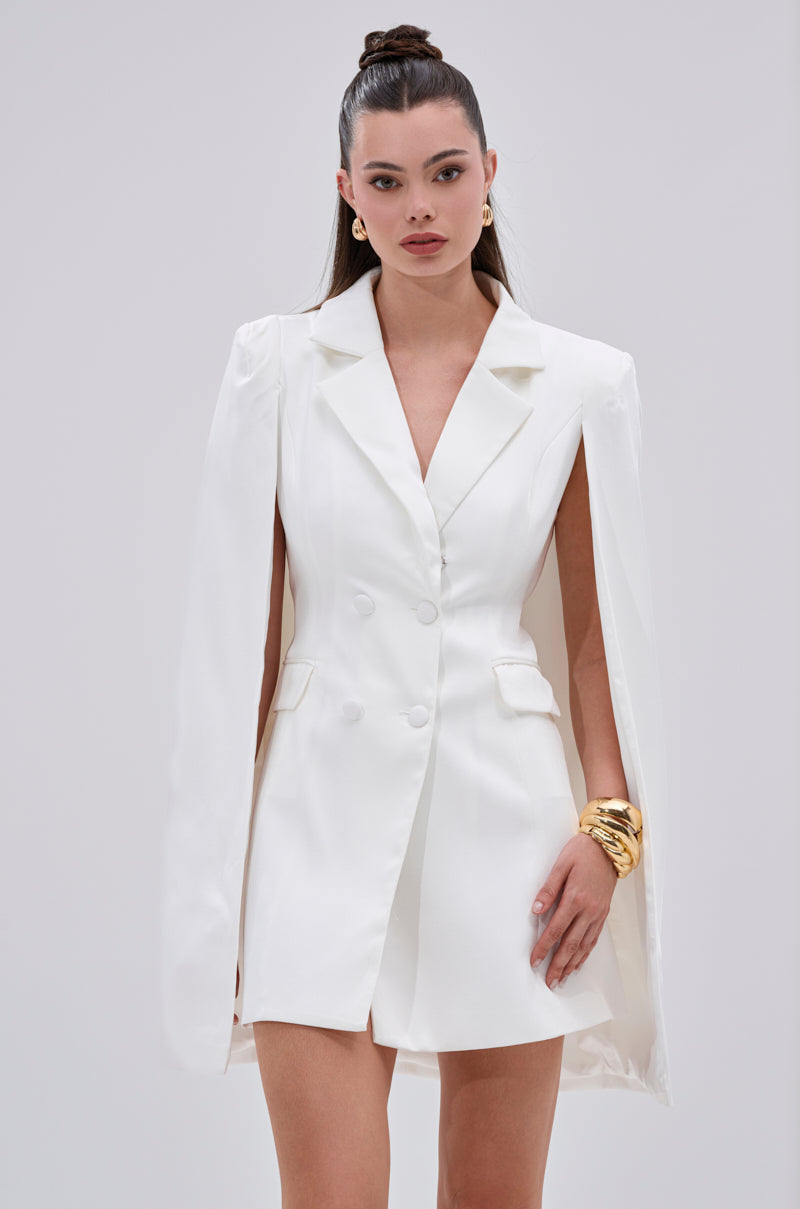 A woman models the A LA MODE MINI BLAZER DRESS IN WHITE, featuring cape sleeves and a bodycon fit. She styles her hair in a high bun and accessorizes with gold earrings and chunky bracelets against a plain light background.