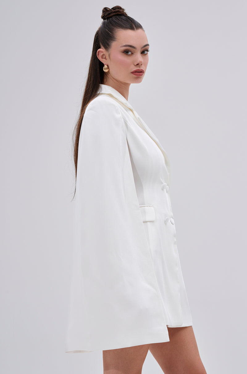 A woman with long brown hair styled half-up wears the A LA MODE MINI BLAZER DRESS IN WHITE, featuring a mini silhouette. She stands sideways against a light gray background, accessorized with gold earrings and a neutral expression.