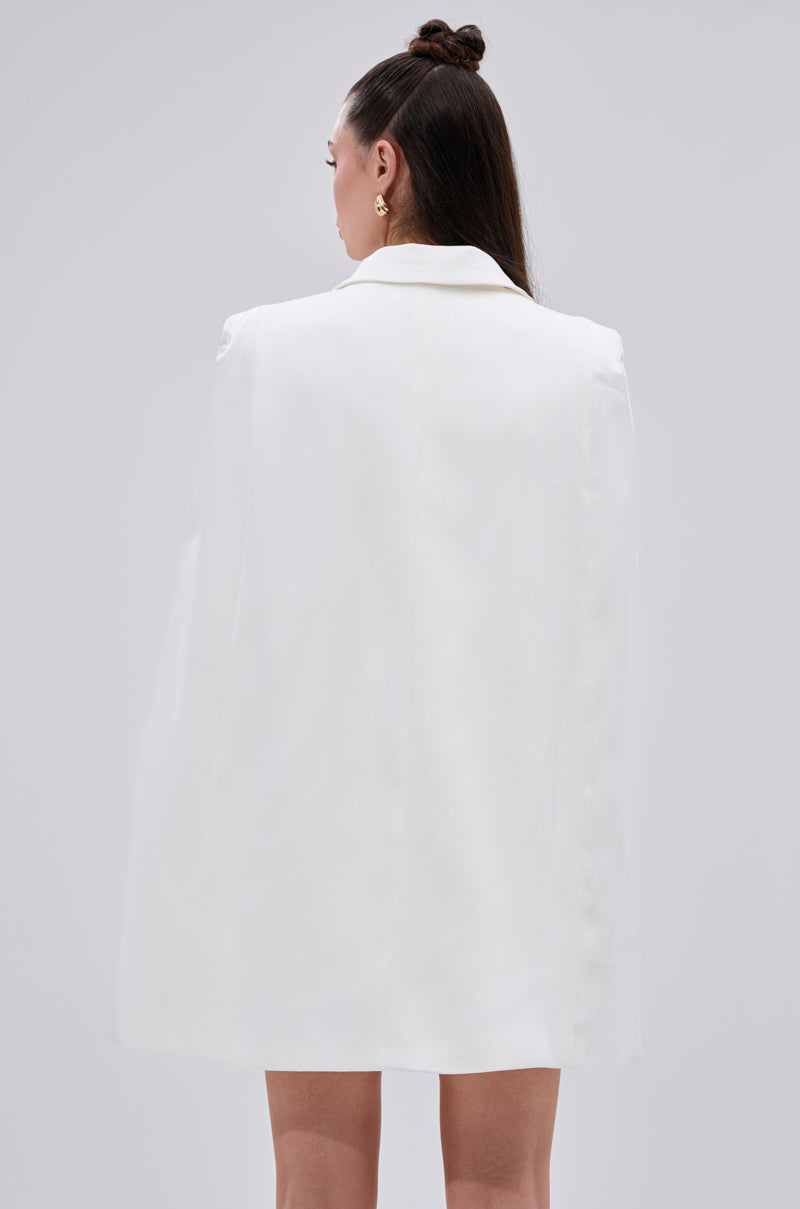 A woman with long dark hair in a half-up bun, facing away, models the A LA MODE MINI BLAZER DRESS IN WHITE—a mini-length cape blazer dress—against a plain light gray background.