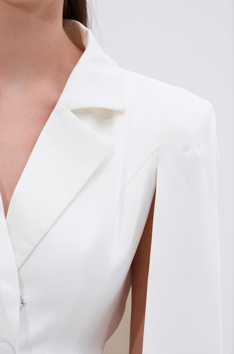 A close-up of a person wearing the A LA MODE MINI BLAZER DRESS IN WHITE, featuring a white cape design, deep V-neckline, and notched lapel, against a plain background.