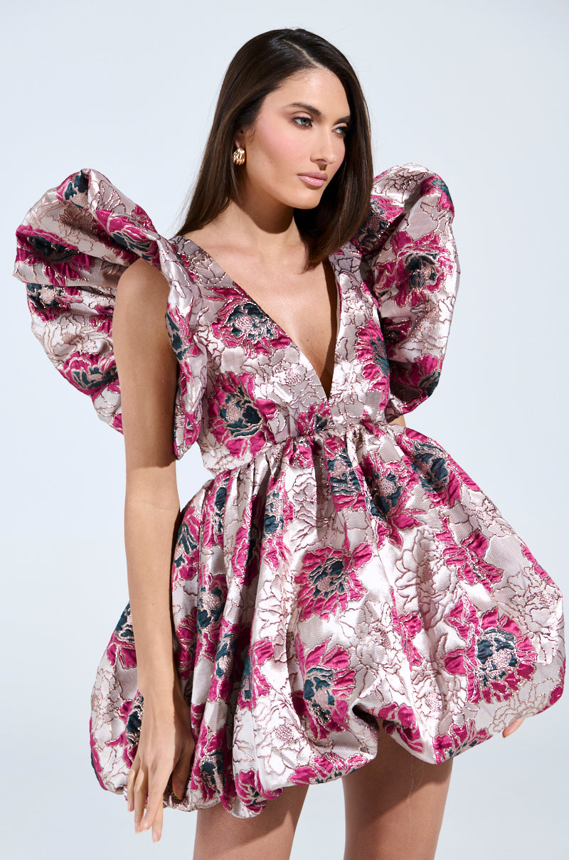 A woman poses confidently in the MAIN CHARACTER ENERGY BROCADE MINI DRESS IN SILVER, featuring a short, floral-patterned design with voluminous ruffled sleeves, shades of pink, purple, and gray, and a deep plunging neckline. Her long brown hair flows against a light background.