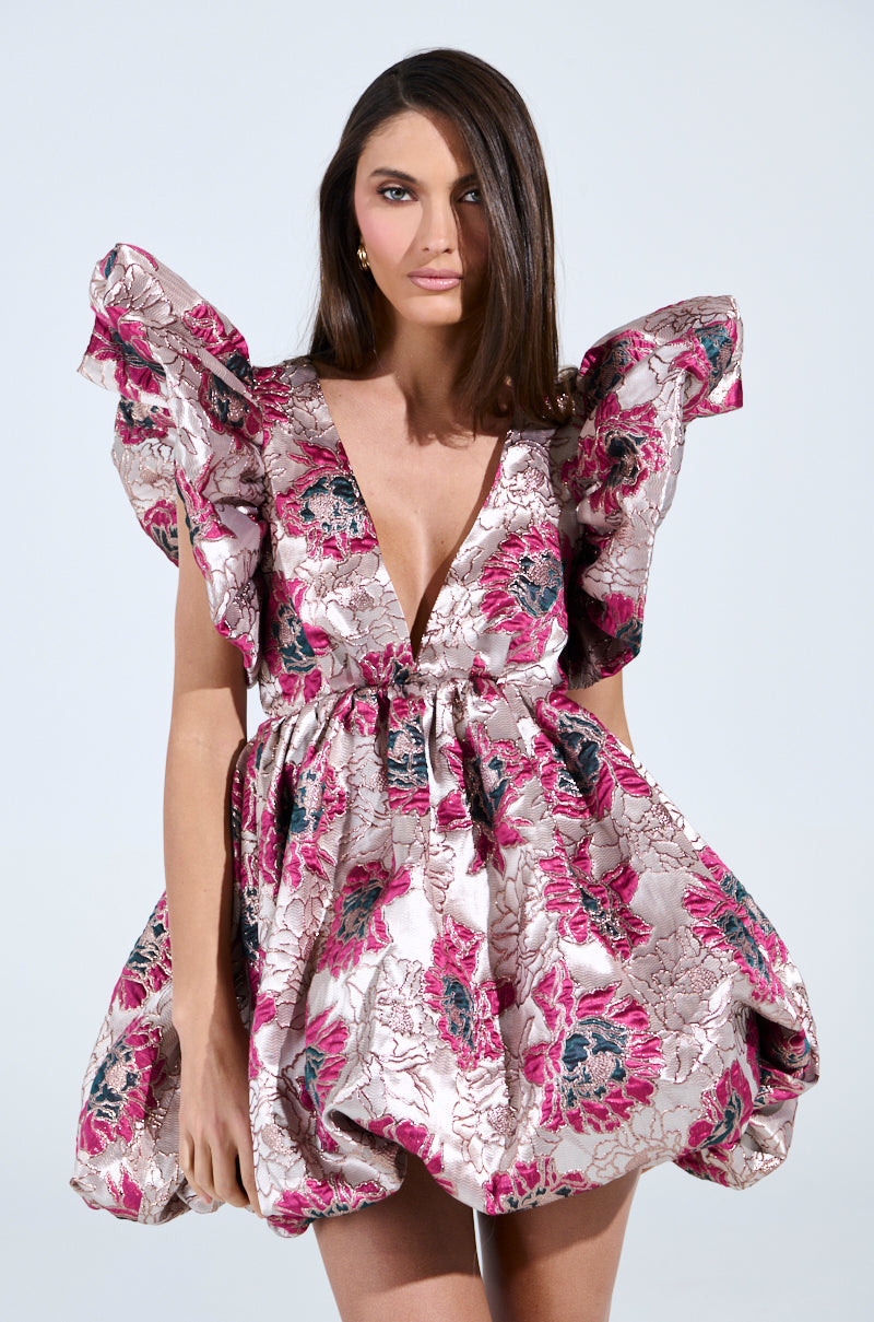A person wearing the Main Character Energy Brocade Mini Dress in Silver, featuring pink, silver, and black flowers with puffed shoulders and a plunging neckline against a plain white background.