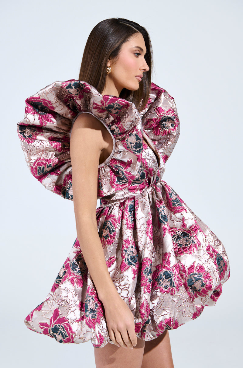 A woman poses elegantly against a plain backdrop in the silver MAIN CHARACTER ENERGY BROCADE MINI DRESS featuring large ruffled shoulders. She accessorizes with gold earrings and her hair is styled straight and sleek.