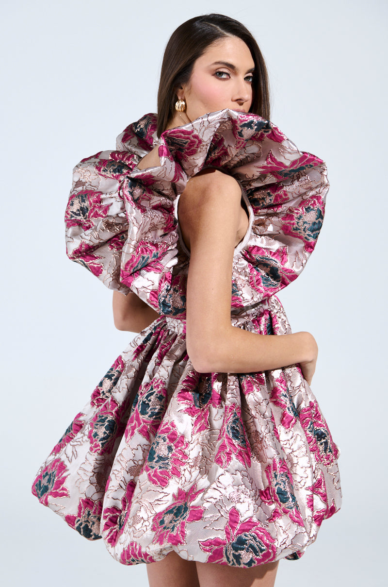 A person in the MAIN CHARACTER ENERGY BROCADE MINI DRESS IN SILVER, adorned with large pink floral patterns, oversized ruffled sleeves, and a voluminous skirt. The metallic fabric glistens as the individual with long dark hair turns towards the camera.