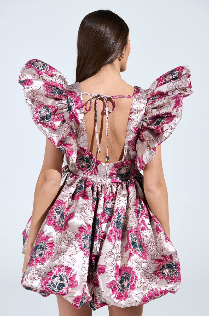 A woman with long brown hair stands facing away, wearing the MAIN CHARACTER ENERGY BROCADE MINI DRESS in a pink and white floral pattern. The dress has large ruffled sleeves, an open back with tie details, and a striking brocade design.