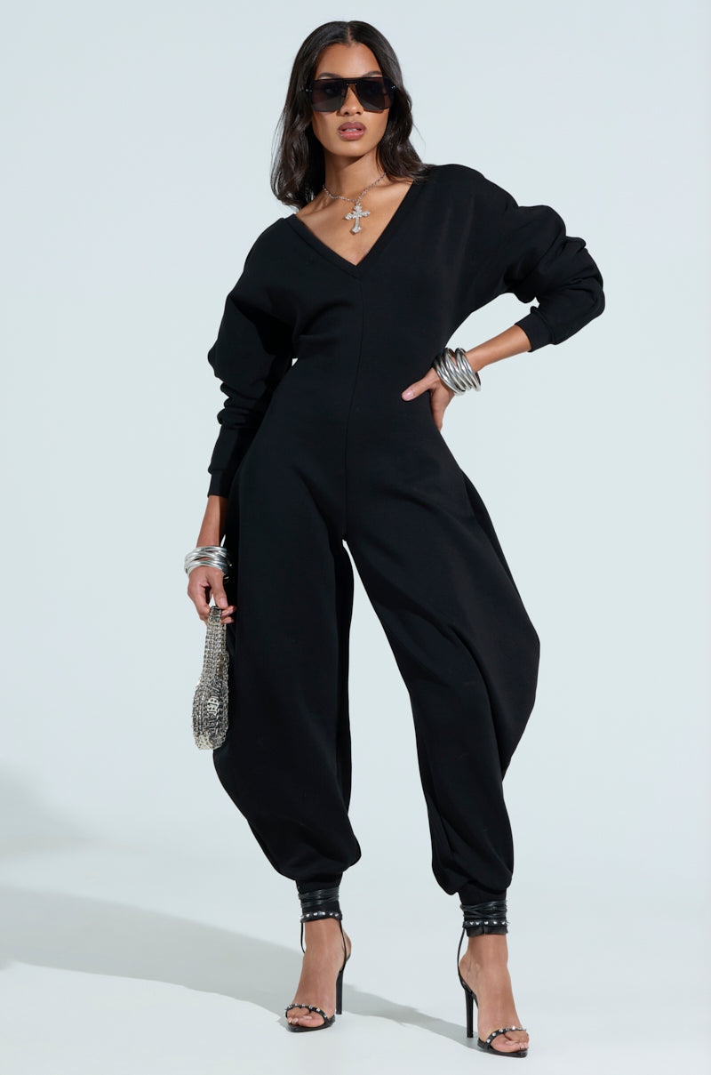 A person poses confidently in a COMFY COZY SWEATSHIRT JUMPSUIT IN BLACK, accessorized with sunglasses, a silver necklace, bracelets, and a sparkly bag. Their look is completed with black high-heeled sandals against a light background.