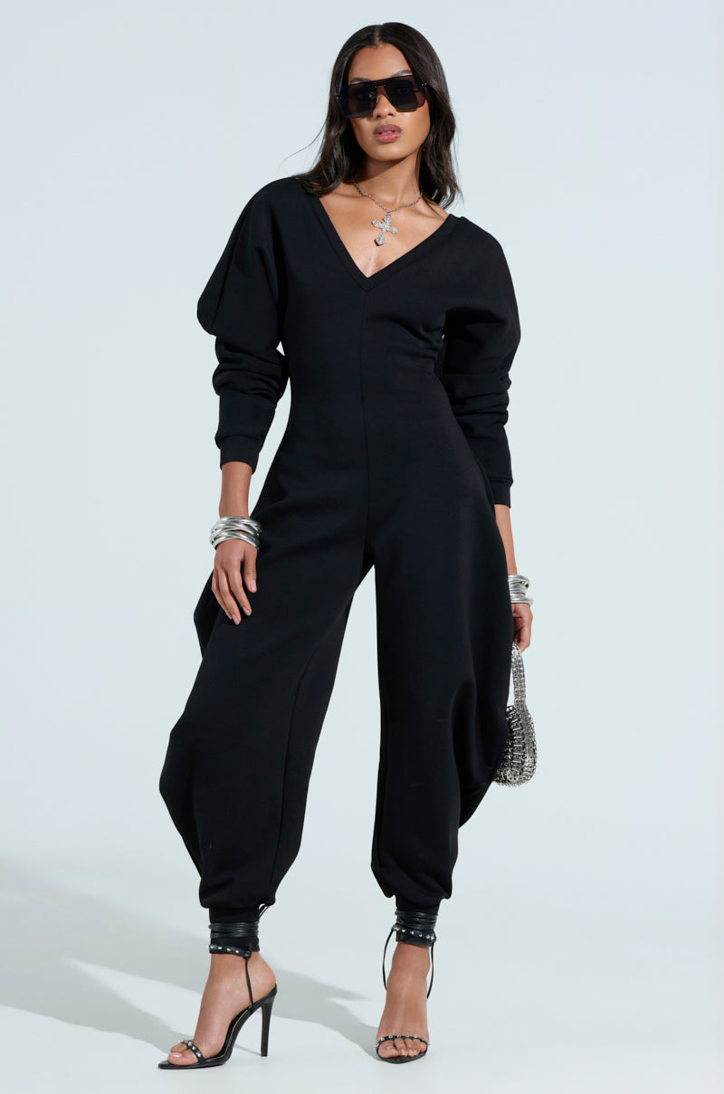 The woman strikes a confident pose in a comfy cozy sweatshirt jumpsuit in black. Accessorized with high-heeled sandals, sunglasses, silver jewelry, and holding a small handbag, her chic style is accentuated against the plain light blue backdrop.
