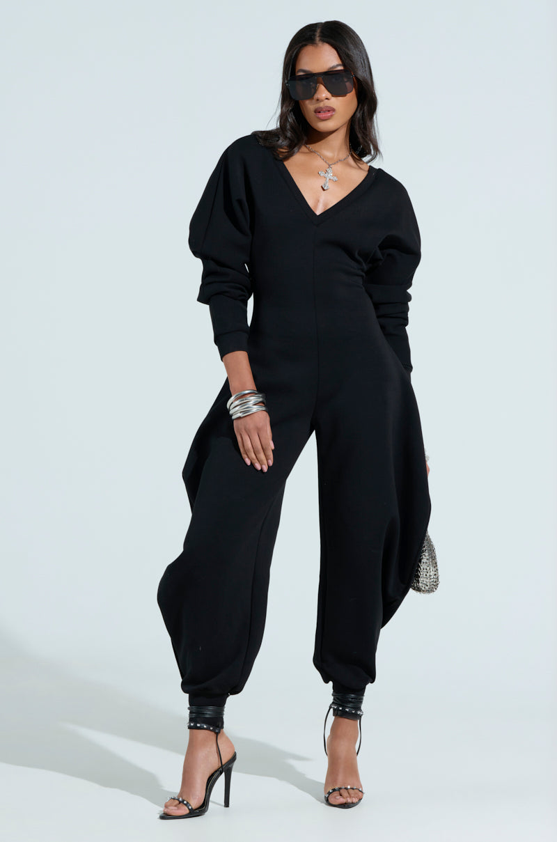 A person in the chic COMFY COZY SWEATSHIRT JUMPSUIT IN BLACK, featuring puffed sleeves and a plunging V-neck, accessorizes with sunglasses, a necklace, bracelets, and high heels. They confidently hold a small bag against a light background.