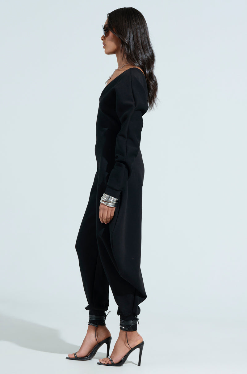 Side profile of a woman in a COMFY COZY SWEATSHIRT JUMPSUIT IN BLACK with a plunging V-neckline and sunglasses. She has long dark hair, multiple bracelets, and stands tall in high heels against a minimalistic light background.