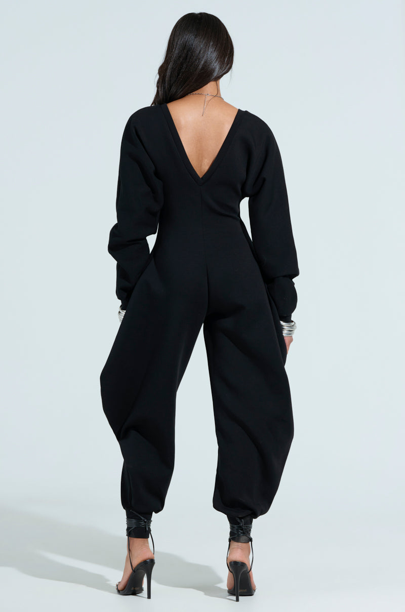 A rear view of someone with long hair wearing the COMFY COZY SWEATSHIRT JUMPSUIT IN BLACK, featuring wide legs and heeled sandals. The jumpsuit contrasts against the plain, light-colored background.