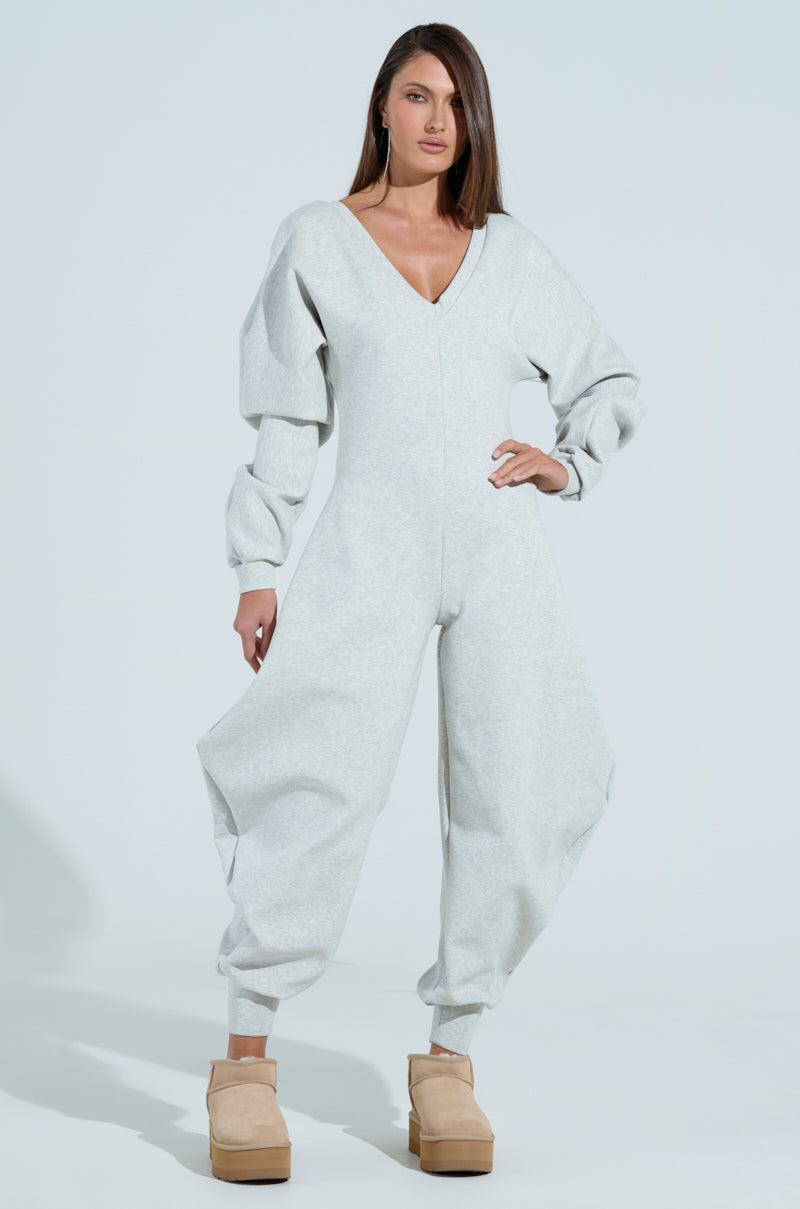 Against a light backdrop, a confident person models the COMFY COZY SWEATSHIRT JUMPSUIT in light gray, showcasing its loose fit with voluminous sleeves and pant legs and a plunging neckline, perfectly paired with beige platform shoes. Long hair cascades gracefully down their back.