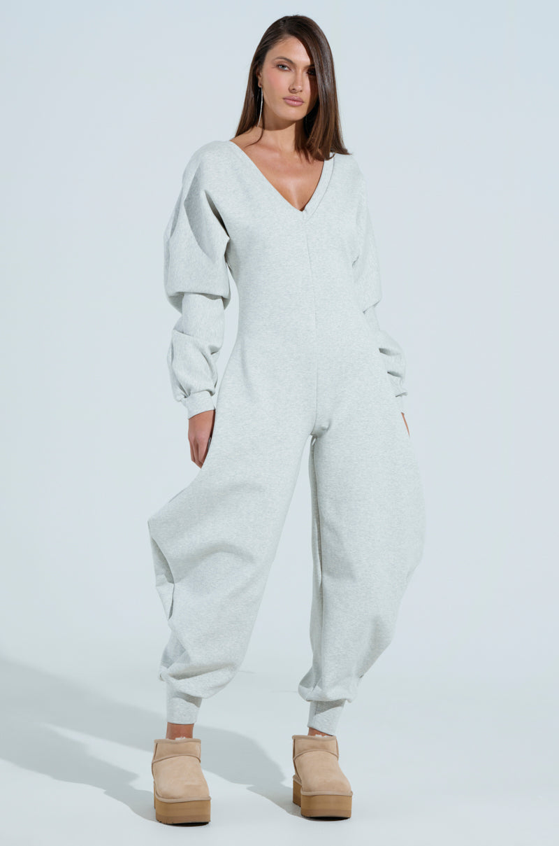A woman poses against a light background in the COMFY COZY SWEATSHIRT JUMPSUIT, light gray with voluminous long sleeves and a plunging neckline. Beige platform shoes complete her look. Her straight hair and confident gaze encapsulate modern elegance.