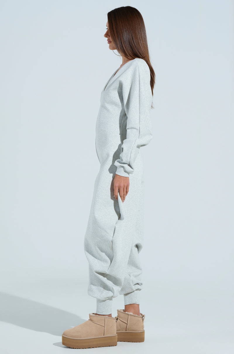 A person with long brown hair stands sideways, wearing a COMFY COZY SWEATSHIRT JUMPSUIT in light gray with elastic ankle hems and tan platform shoes, against a plain light blue background.