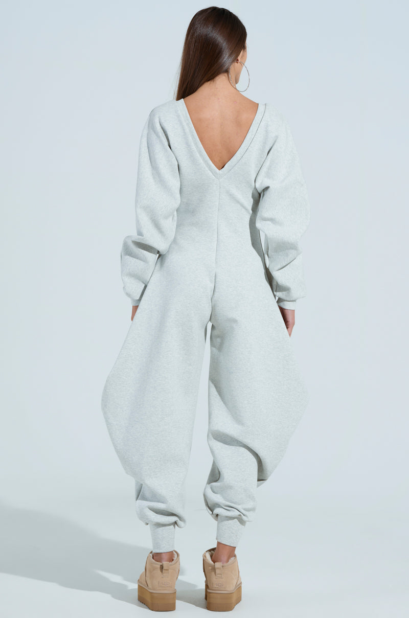 Wearing the COMFY COZY SWEATSHIRT JUMPSUIT, a person stands on platform shoes, facing away against a white background. The jumpsuit features voluminous sleeves and a deep V-neck back, with their long brown hair cascading downward.