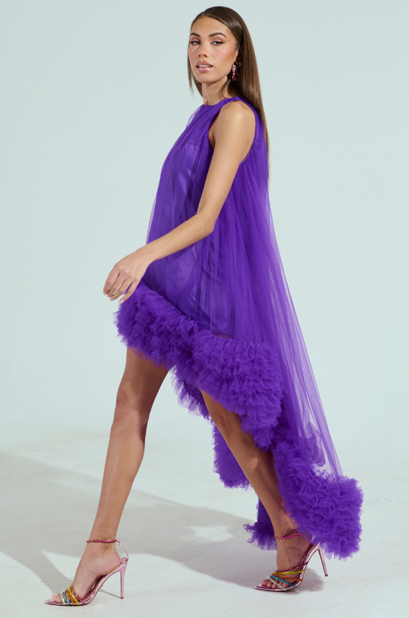 A woman confidently walks in the ARI TULLE RUFFLE HIGH LOW DRESS IN PURPLE, featuring a flowy silhouette and tulle ruffle trim. Her metallic strappy heels perfectly complement the outfit against a light-colored, plain background.