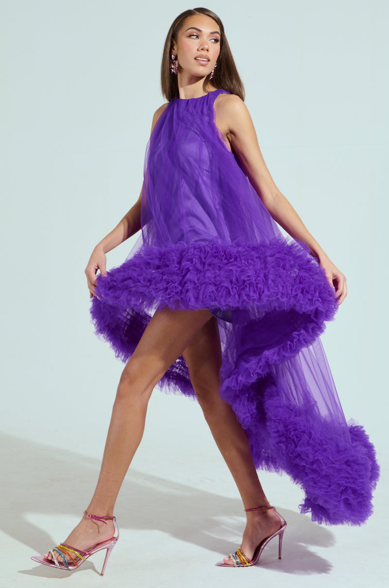 The model confidently walks in the ARI TULLE RUFFLE HIGH LOW DRESS IN PURPLE, featuring a captivating tulle ruffle hem and sleeveless sheer fabric. She pairs it with pink high heels and statement earrings, set against a light background that highlights her elegant ensemble.
