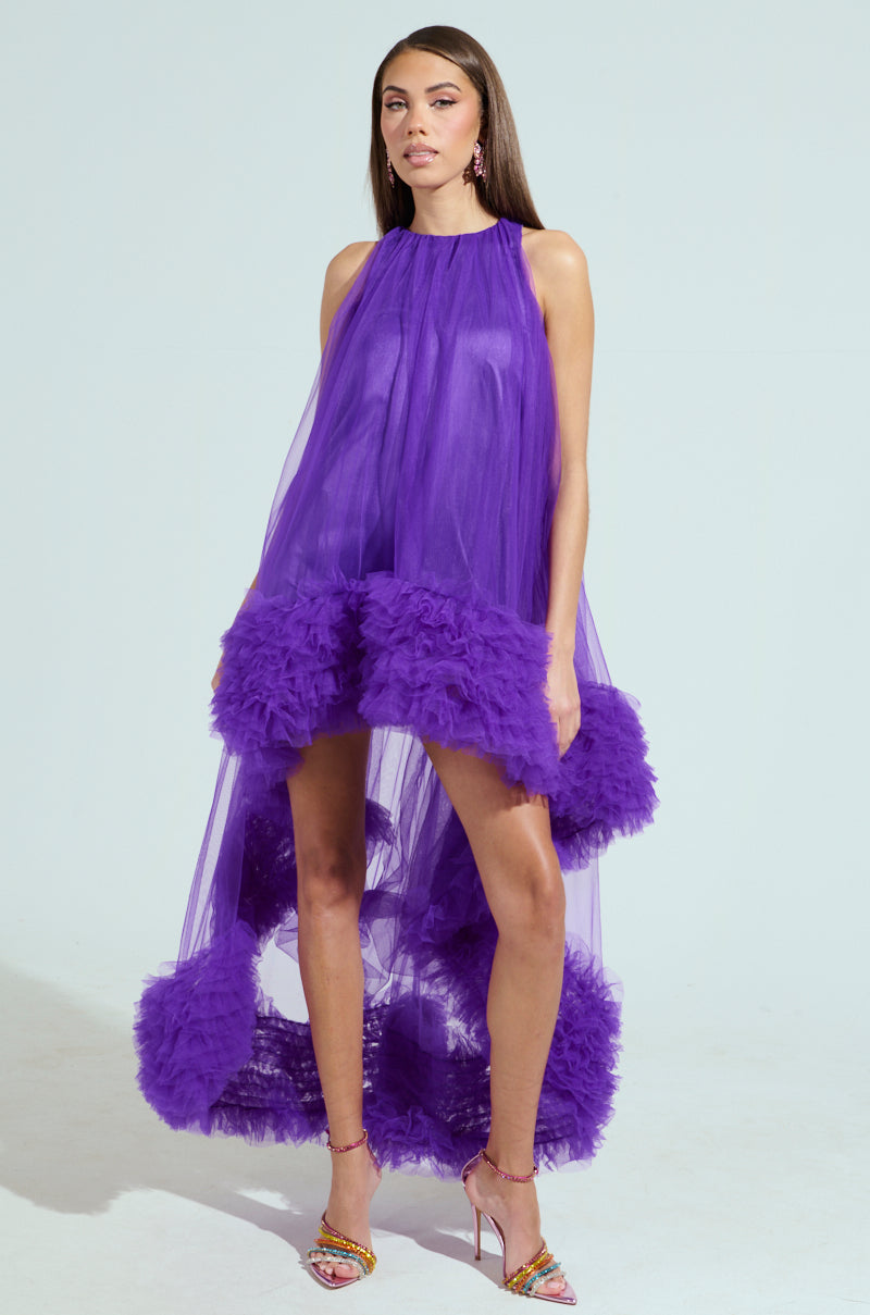 Wearing the ARI TULLE RUFFLE HIGH LOW DRESS IN PURPLE, a woman stands confidently against a light backdrop. Her long hair, purple earrings, and colorful high heels complete her elegant and stylish look.