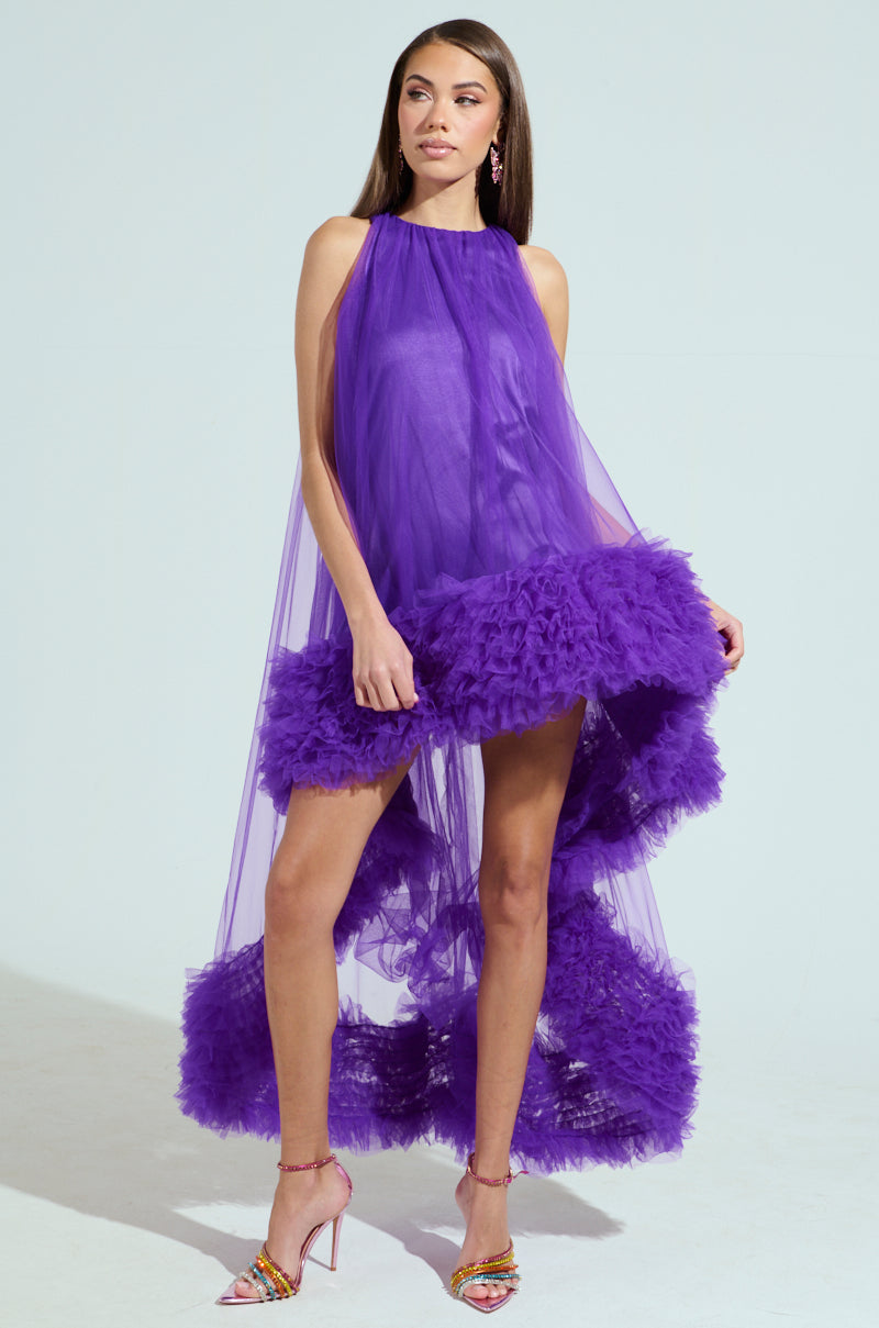 A woman confidently poses in the ARI TULLE RUFFLE HIGH LOW DRESS IN PURPLE. Her long brown hair cascades over the tulle ruffle gown as she stylishly stands on multicolored heels against a light background.