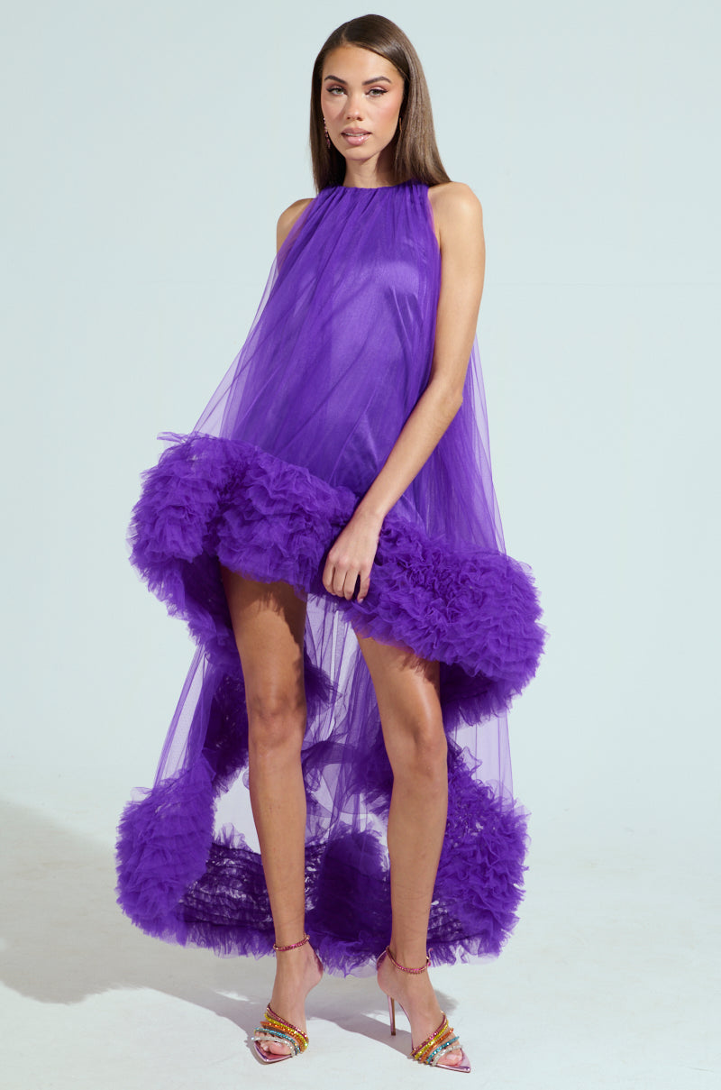 A woman models the ARI TULLE RUFFLE HIGH LOW DRESS IN PURPLE, featuring a vibrant purple hue, tulle ruffle hem, and high neck. The dress flows elegantly to the floor as she exudes confidence against a plain background, paired with colorful high-heeled sandals.