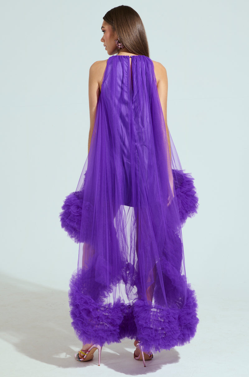 A woman poses against a light background, wearing the ARI TULLE RUFFLE HIGH LOW DRESS IN PURPLE. Her back faces the camera, revealing a stunning dress with sheer fabric and fluffy tulle ruffles at the bottom, while her long brown hair cascades down.